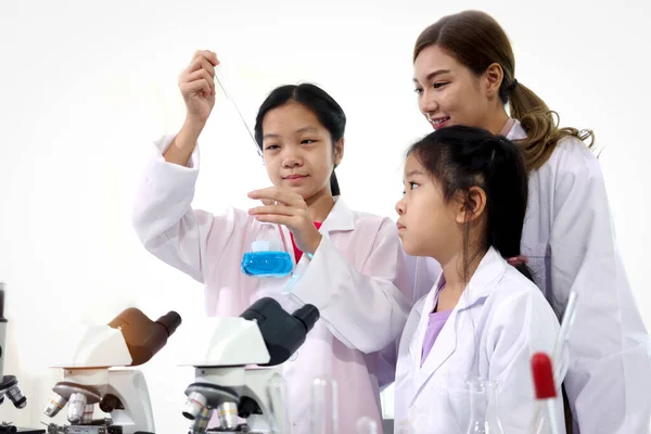 Students and teacher in lab coat have fun together while learn science experiment in laboratory. Young adorable Asian scientist kid adding some chemical to blue flask with pipette, little children students study research at school lab. Science educat