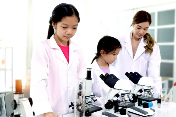 Students and teacher in lab coat have fun together while learn science experiment in laboratory. Young adorable Asian scientist kid using microscope, apparatus and lab equipment for research. Science education for kid concept.
