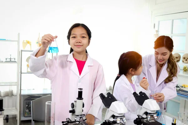 Students and teacher in lab coat have fun together while learn science experiment in laboratory. Young adorable Asian scientist kid showing blue flask with teacher and classmate as blur background, little children students study research at school la