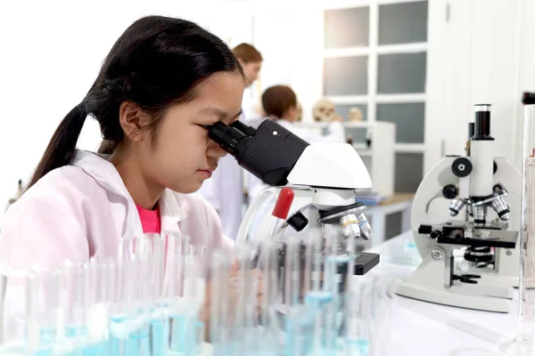 Adorable schoolgirl in lab coat doing science experiments, young scientist looking through microscope and learn science experiment in laboratory. Asian kid using apparatus and lab equipment for research with teacher and classmate as blur background.