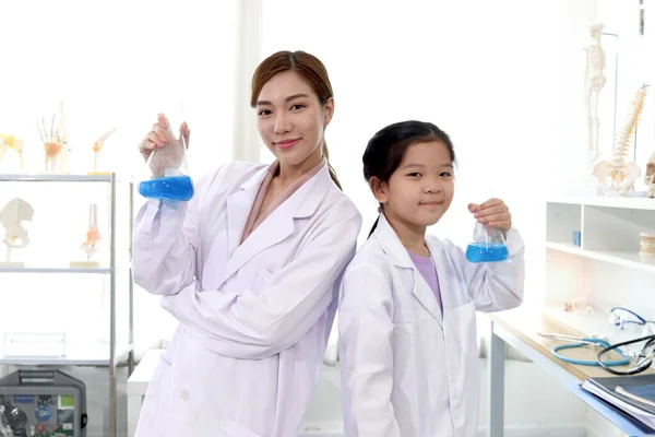 Portrait of beautiful Asian science teacher and adorable schoolgirl in lab coat holding blue chemical flasks and standing back to back with arms crossed in laboratory. Smiling young scientist with kid have fun together and doing science experiments,
