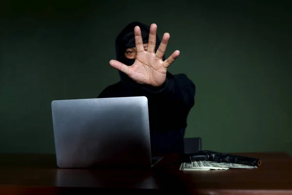 Criminal man in black hood with laptop computer, a gun and money at the at desk  reaching out his hand to camera , mask thief robber committing committed cyber crimes online.
