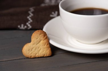 Kurabiyeler beyaz bir fincan tabağın ve kahverengi ahşap bir fotografın üzerinde bir fincan sade kahvenin yanında yakın plan kalp şeklinde.