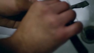 Lavaboda akan suyun altında tıraş bıçağının kıllarını yıkamak. Çerçevede, erkeklerin elleri suyun altında parlak bir bıçağı temizlerken görülüyor..