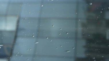 Çerçeve penceresinin mavi camında dikey yağmur damlası görüntüsü. Çerçevenin altında küçük bir bina silueti görülebilir..