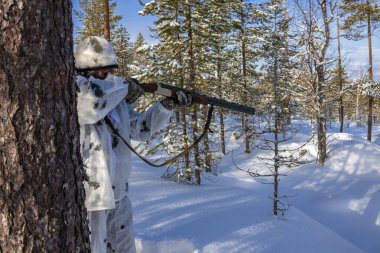 Avcı kışın kamuflajlı, yüzü maskeyle kaplı ve ateş etmeye hazırlanıyor. Avcı maskeli adam karlı ormanda tüfekle nişan alır.. 
