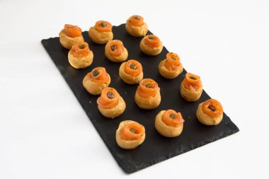 Küçük, lezzetli, iştah açıcı, ısırık büyüklüğünde, tütsülenmiş somon ve kapari kano profiterolleri, siyah dikdörtgen tepside kekler.