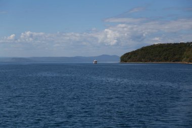 Deniz manzarası, yeşil adalar, pelerinler ve arkasında güzel bulutlar ve mavi bir gökyüzü olan beyaz lüks bir yat. Rusya 'nın Vladivostok kentindeki Russky adasındaki Novik limanı