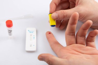 Bir elde iki el, bir mızrakla parmak ve boş bir hızlı antojen ekspres Coronavirus covid-19 pandemik test örneğinden kan örneği alarak beyaz bir arkaplanda izole edilmiş.