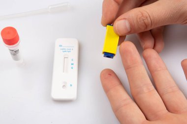 Bir elde iki el, bir mızrakla parmak ve boş bir hızlı antojen ekspres Coronavirus covid-19 pandemik test örneğinden kan örneği alarak beyaz bir arkaplanda izole edilmiş.