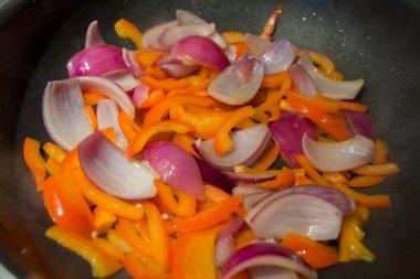 Doğranmış sebze dilimlerinin, kırmızı dolma biberlerin ve kızarmış tavada pişen mor soğanların üst görüntüsünü kapat.