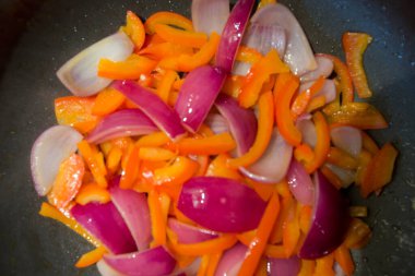 Doğranmış sebze dilimlerinin, kırmızı dolma biberlerin ve kızarmış tavada pişen mor soğanların üst görüntüsünü kapat.