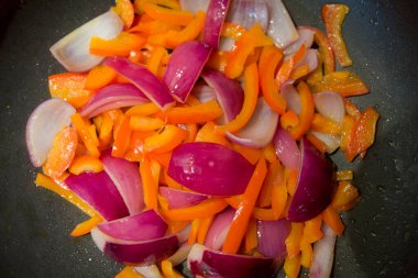 Doğranmış sebze dilimlerinin, kırmızı dolma biberlerin ve kızarmış tavada pişen mor soğanların üst görüntüsünü kapat.