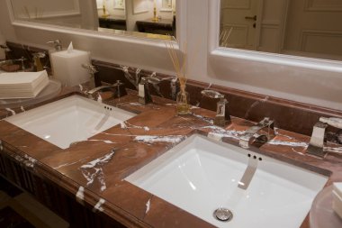 Kahverengi bej rengi granit mermer tezgahlı lüks bir banyo üst tahtası, iki beyaz seramik lavabo, büyük dikdörtgen aynalar, klasik duvar lambası, parfüm yağı çubukları, kağıt havlular.