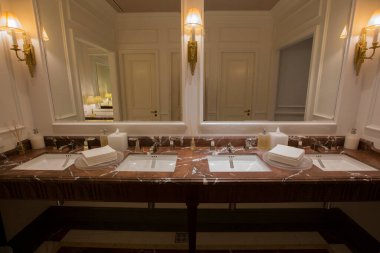 Kahverengi bej rengi granit mermer tezgahlı lüks bir banyo üst tahtası, iki beyaz seramik lavabo, büyük dikdörtgen aynalar, klasik duvar lambası, parfüm yağı çubukları, kağıt havlular.