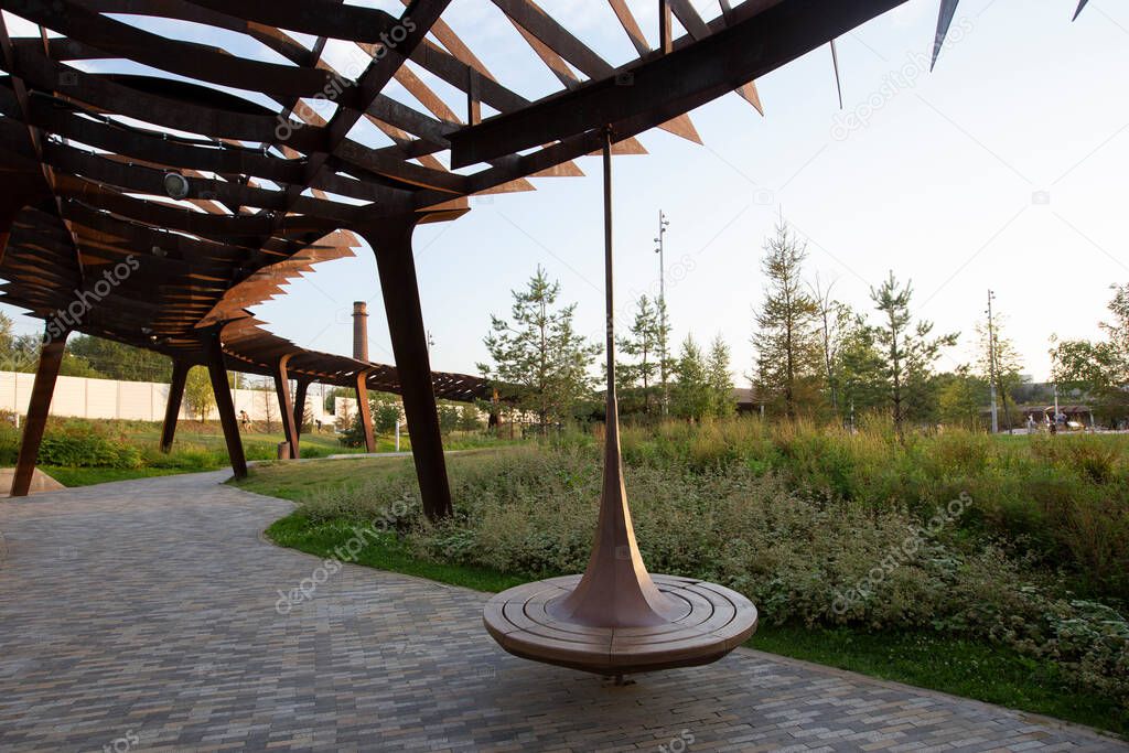 spinning wooden bench and metal pergola. Walkway with grass lawn, bushes, plants, trees in background. Tyufeleva Roshcha city park, Zilart, Moscow, Russia. !melk landscape architecture urban design