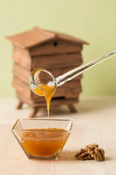 透明ガラスのスプーンで茶色のオレンジの蜂蜜を皿のボウルに注ぎ クルミと緑の背景の後ろに小さな木造の小屋のモデル — ストック写真