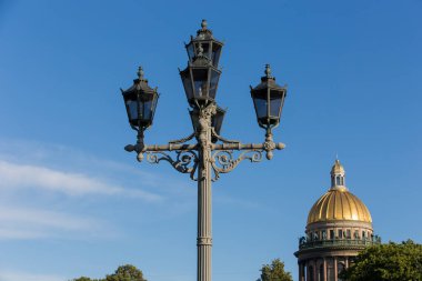 SAINT PETERSBURG, RUSSIA 13.08.2021 Classic candelabra street light with five lamps. Saint Isaac's Cathedral's golden cupola dome, granite columns, statues, green trees foliage on blue sky background
