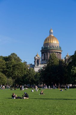 SAINT PETERSBURG, RUSSIA 13.08.2021 people resting on the grass in Alexander Garden park on sunny summer day with green trees foliage and St. Isaac's Cathedral golden cupola dome in the back 