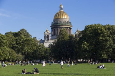 SAINT PETERSBURG, RUSSIA 13.08.2021 people resting on the grass in Alexander Garden park on sunny summer day with green trees foliage and St. Isaac's Cathedral golden cupola dome in the back 