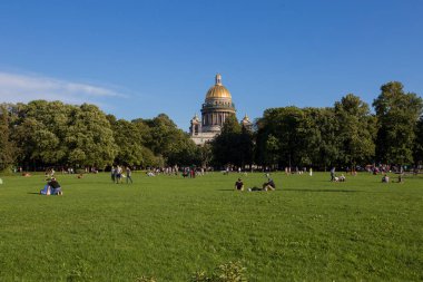 SAINT PETERSBURG, RUSSIA 13.08.2021 people resting on the grass in Alexander Garden park on sunny summer day with green trees foliage and St. Isaac's Cathedral golden cupola dome in the back 