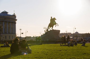 SAINT PETERSBURG, RUSSIA 13.08.2021 contrast shot of people sitting on grass in Alexander Garden park with silhouette of Bronze Horseman sculpture of Peter the Great on blue sky and sun background