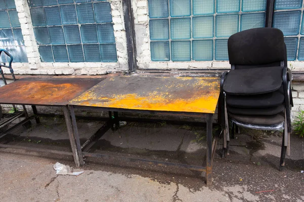Eski, tozlu ofis sandalyeleri ve paslı, sarı metalik bir masa dışarıda duruyor. Sovyet mavisi bir camın üzerinde. Pencereler de arka planda.