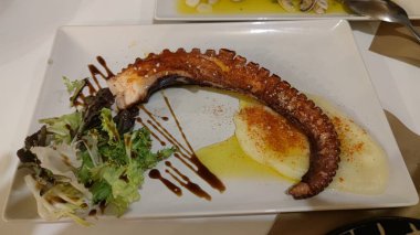 Izgara ahtapot bacağı, patates püresi ve beyaz tabakta yeşil salata.