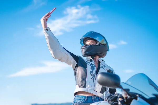 革のジャケットに身を包んだ若い女性バイカーと安全ヘルメットは彼女の道路のオートバイに彼女のスマートフォンで自撮りを取ります — ストック写真