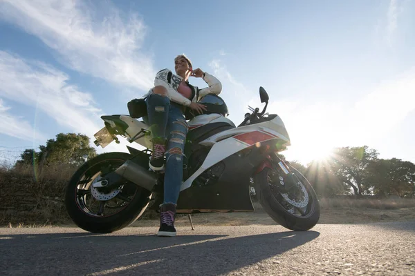 バイクの装備を着た若い可愛い女が道路のバイクにもたれて笑ってた 背景の夕日は — ストック写真