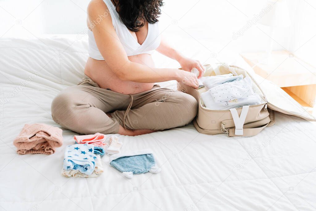 young pregnant woman sitting on the bed with the clothes of her future baby preparing the suitcase for the hospital.
