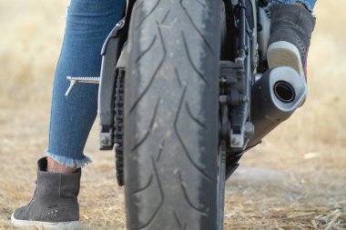 Motosikletin tekerleğinin dikiz manzarası. Motosiklet sürmek için güvenlik botu ve korunaklı kot giyen bir motorcu kadının ayakları.