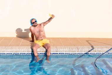 smiling young hipster man with beard, yellow swimsuit and sunglasses takes a selfie with his smartphone in the pool with his feet in the water while listening to music with his blue headphones.