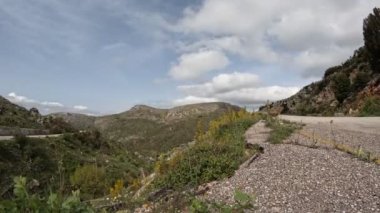 Time lapse video of a rural road with flowers moving at it's side and clouds moving at the background.