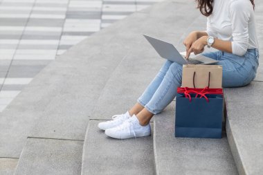 Genç bir kadın alışverişe çıktıktan sonra bir mağazanın önünde dizüstü bilgisayardan işlerini kontrol ediyor. Yeni yıl arifesinde uygun fiyatlara harika ürünler almak için alışveriş fikirleri..