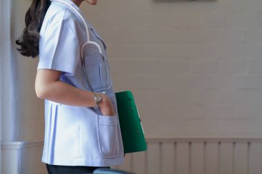 Üniformalı bir kadın doktor, elinde hastanın muayene ve teşhisini gösteren bir pano olan pencerenin yanında duruyor..