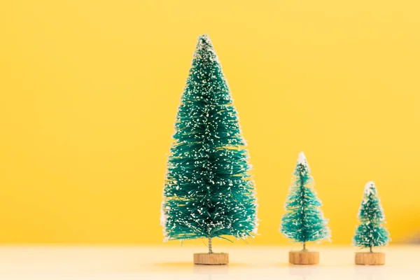 miniature green Christmas tree sits on white table and has bright yellow background that contrasts nicely with green Christmas tree and has a copy space to insert text to create greeting card.
