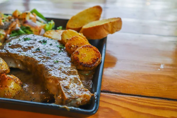 Grilled beef steak with gravy is served with a fruit and vegetable salad inside a black ceramic plate on the dining room table to prepare the steak for a festive dinner.