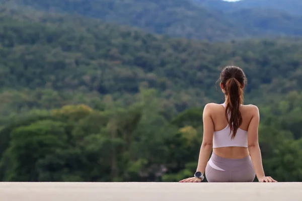long haired woman in sports bra sits alone on roadside on dam crest against blurred background of green nature alone after jogging.  young woman sits alone on blurry background and has Copy Space