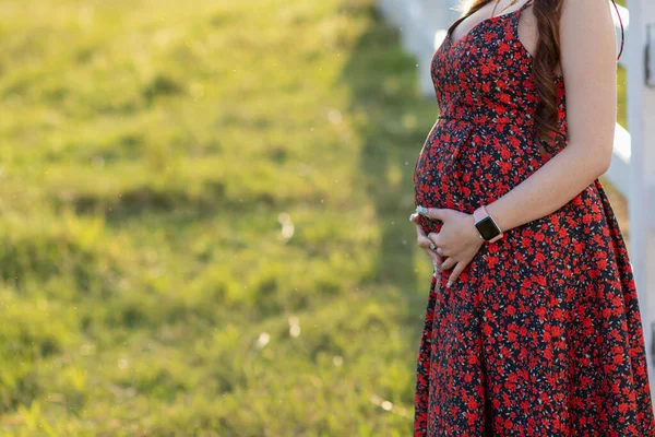 A pregnant woman nearing birth, wearing a vintage floral red maternity dress, is standing and grabs her stomach to relax in the grass and breathe in the fresh air, giving the unborn child good air.