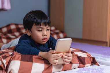 Tatlı Asyalı çocuk akıllı telefon kullanarak yatağında uzanıyordu. Evde boş zamanlarında iyi eğlenceler. Virüs havalandırması, online teknoloji