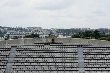 Turuncu gömlekli bir adam boş bir stadyum tribününün başında duruyor ve Paris şehrine bakıyor.