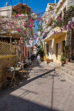 Aegina Adası Yunanistan - 4.27.2022: Dükkanları olan sokak, dar kasaba sokaklarında koruyucu maske takan yürüyen yaşlı kadın. Kapalı dükkanlar, yaz ışığı. Asılı çiçek süslemesi