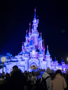Paris, Fransa - 04 / 05 / 2022: Geceleri Uyuyan Güzel Kalesi. İnsanlar Disneyland Parkları 'nın ünlü ve simgesel binasına yürüyorlar. Mor ışıklar kaleyi aydınlatıyor..