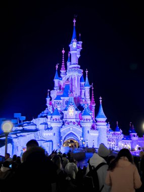 Paris, Fransa - 04 / 05 / 2022: Geceleri Uyuyan Güzel Kalesi. İnsanlar Disneyland Parkları 'nın ünlü ve simgesel binasına yürüyorlar. Mor ışıklar kaleyi aydınlatıyor..