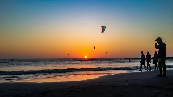 Kite surfing at sunset. Kite boarding outdoor sport activity. People silhouette walking on sandy beach at sunrise with orange sun and colorful beautiful sky. Kite surfers flying in freestyle