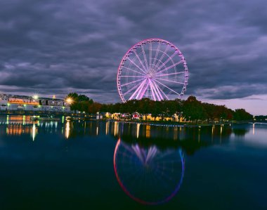 Alacakaranlık mavi saatinde Old Port 'ta Montreal Grand Ferris Wheel' in uzun pozu. Arka plan bulutlu, karamsar bir gökyüzü. Önplan, sonbahar renk ağaçları ve Saint Lawrence (Laurent) nehrinin yansımasıdır..