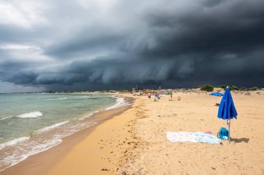 Sicilya 'da güzel güneşli bir sahil. Önünde mavi bir şemsiye ve siyah ve fırtınalı bir gökyüzü var.
