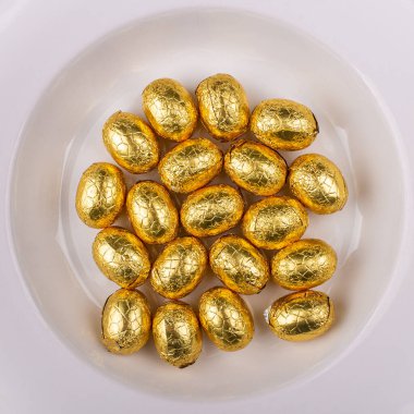 Top view of a pile of yellow golden, wrapped in shiny tinfoil, chocolate shiny Easter eggs in a white round bowl