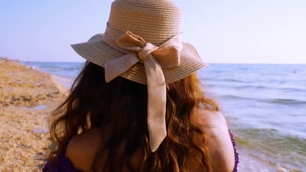 一个戴着一顶帽子的棕色头发的女孩坐在沙滩上 看着大海 调整她的头发 暑假和旅行的概念 — 图库视频影像